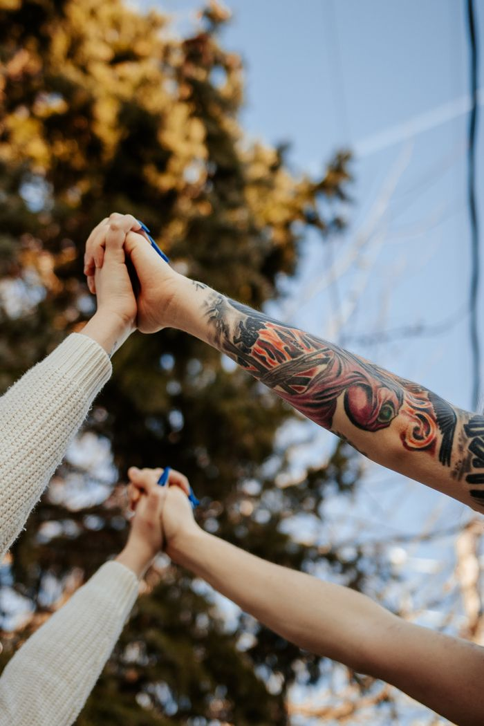 A close-up of hands with tattoos and knit sleeves held together against a blue sky, symbolizing unity and connection.